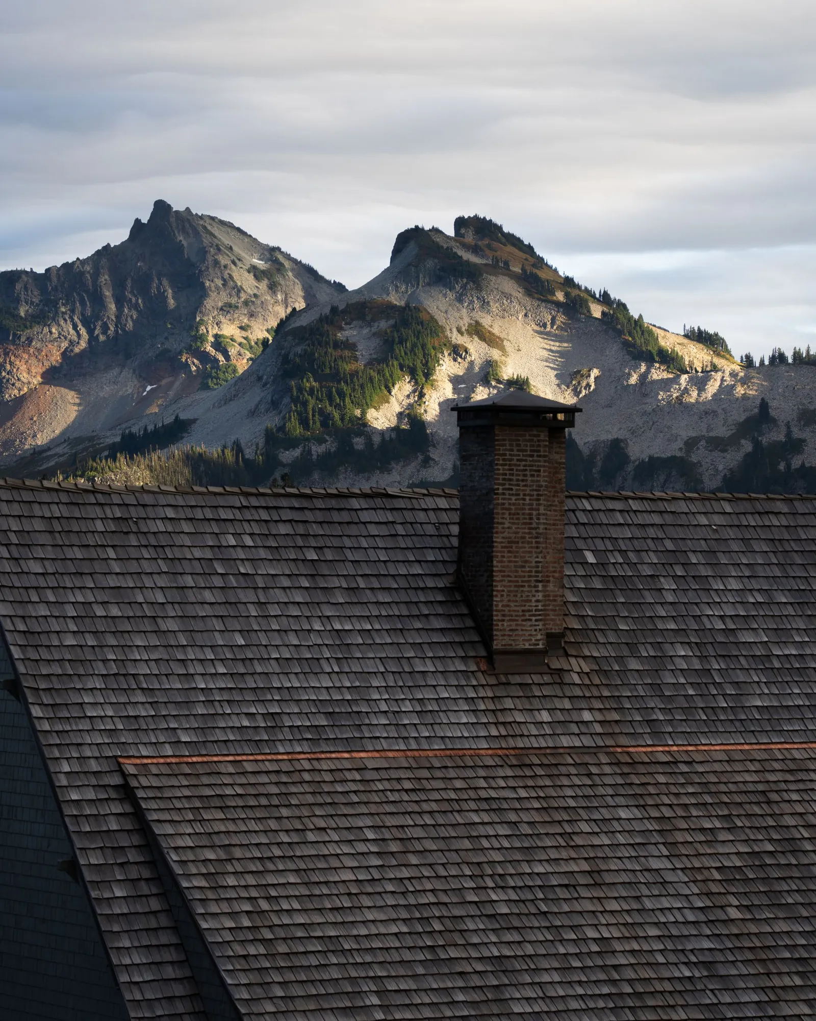 Brick chimney rising from a shingled roof with rugged mountains behind