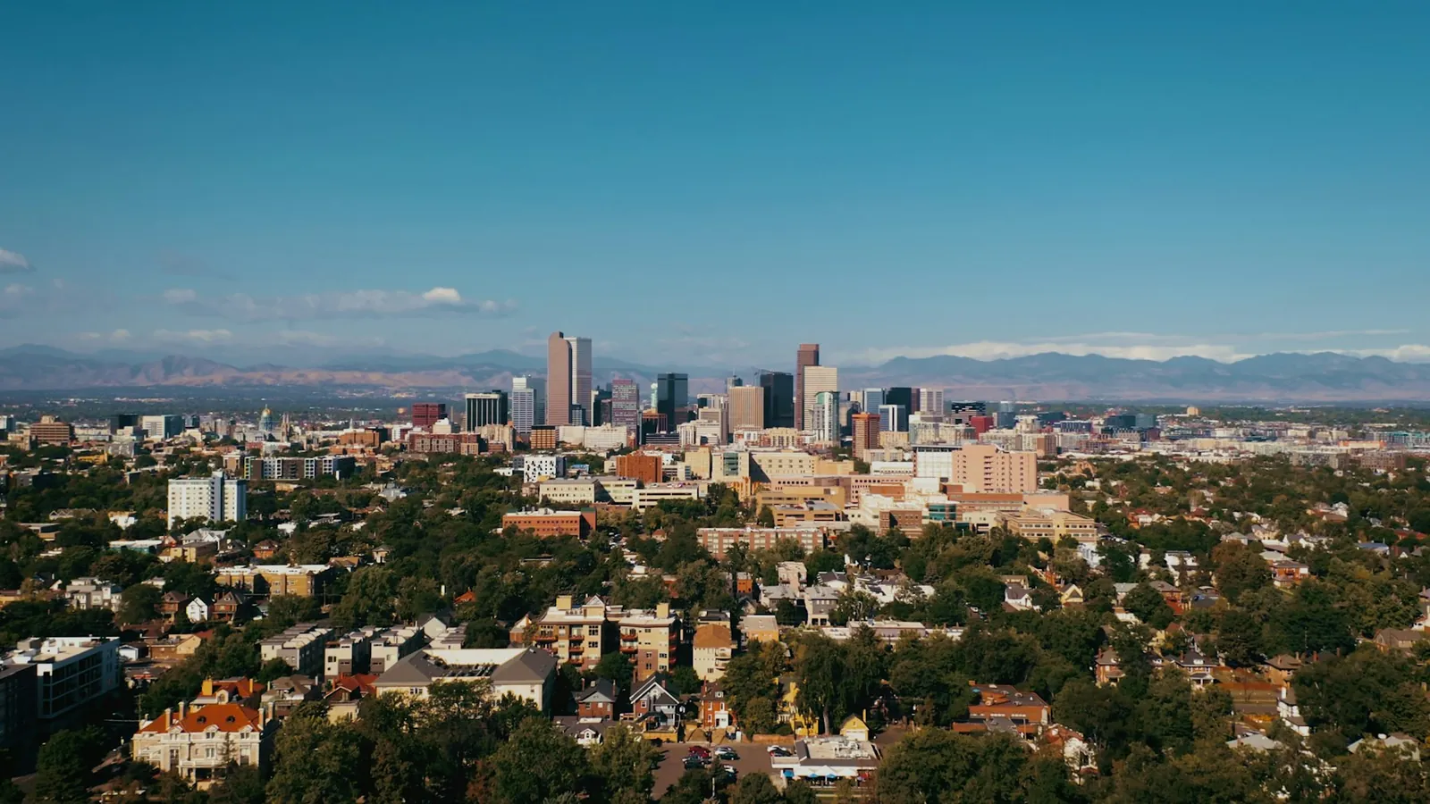 Downtown Denver skyline above tree-lined neighborhoods with the Rocky Mountains beyond